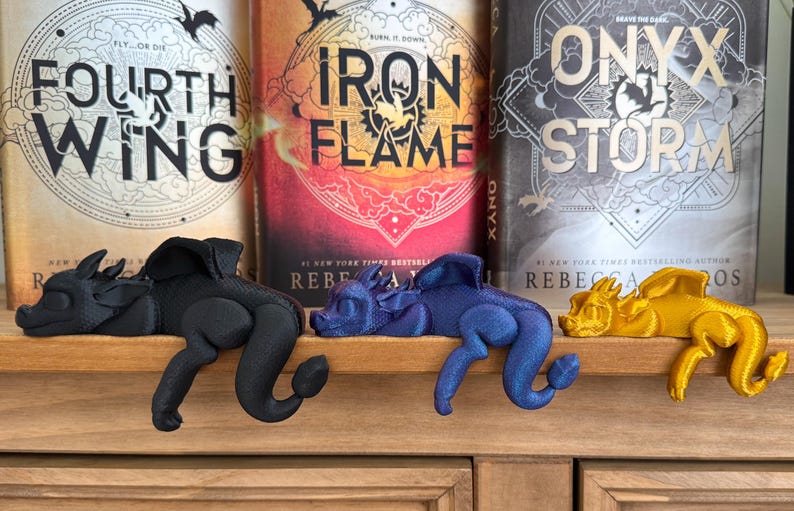 May include: Three 3D printed dragon bookends in black, blue, and gold. The bookends are resting on a wooden shelf in front of three fantasy novels titled "Fourth Wing", "Iron Flame", and "Onyx Storm".