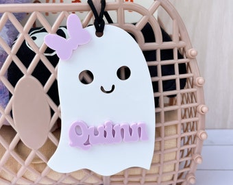 May include: A white ghost-shaped tag with the name "Quinn" in lavender letters, decorated with a lavender butterfly. The tag is suspended from a black cord attached to a pink basket containing items, including a book and a plush toy.