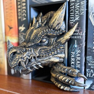 May include: A gold and black dragon head bookend with a textured finish. The dragon's mouth is open, revealing sharp teeth. The bookend is holding two books, one of which is titled "When the Moon Hatched".