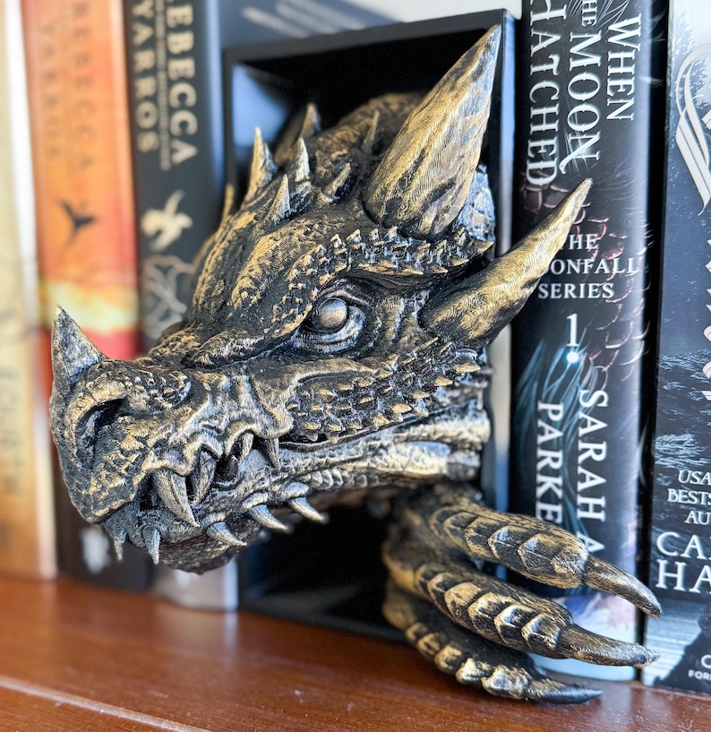 May include: A gold and black dragon head bookend with a textured finish. The dragon's mouth is open, revealing sharp teeth. The bookend is holding two books, one of which is titled "When the Moon Hatched".