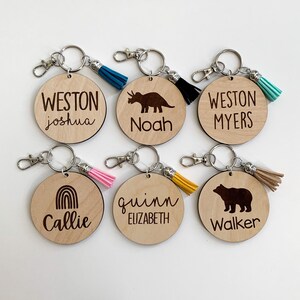 May include: Six wooden keychains with different designs and names. The keychains feature a dinosaur, a rainbow, a bear, and the names "Weston Joshua", "Noah", "Weston Myers", "Callie", "Quinn Elizabeth", and "Walker".