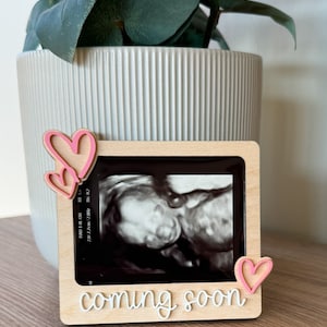 May include: A wooden picture frame with a sonogram image, decorated with pink hearts. The frame has the words "coming soon" in a cursive font. The frame is displayed in front of a white pot with a green plant.