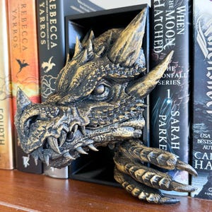 May include: A gold dragon bookend with a detailed, textured design. The dragon's head is facing forward with its mouth open, revealing sharp teeth. The dragon's wings are spread out, and its claws are curled. The bookend is sitting on a shelf with books behind it.