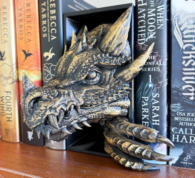 May include: A gold dragon bookend with a detailed, textured design. The dragon's head is facing forward with its mouth open, revealing sharp teeth. The dragon's wings are spread out, and its claws are curled. The bookend is sitting on a shelf with books behind it.