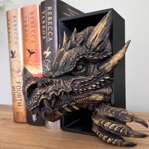 May include: A gold dragon head bookend with a black frame. The dragon has a textured, scaly surface and a fierce expression. The bookend is sitting on a wooden shelf with three books.