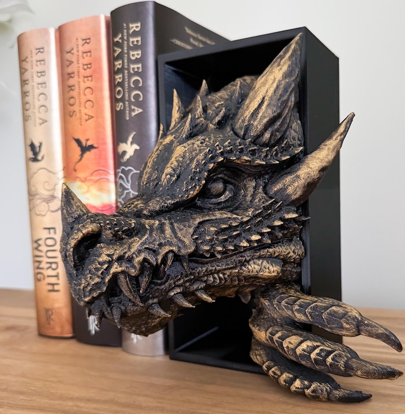 May include: A gold dragon head bookend with a black frame. The dragon has a textured, scaly surface and a fierce expression. The bookend is sitting on a wooden shelf with three books.