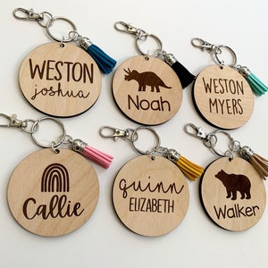 May include: Six wooden keychains with different designs and names. The keychains feature a dinosaur, a rainbow, a bear, and the names Weston Joshua, Noah, Weston Myers, Callie, Quinn Elizabeth, and Walker. Each keychain has a small tassel.