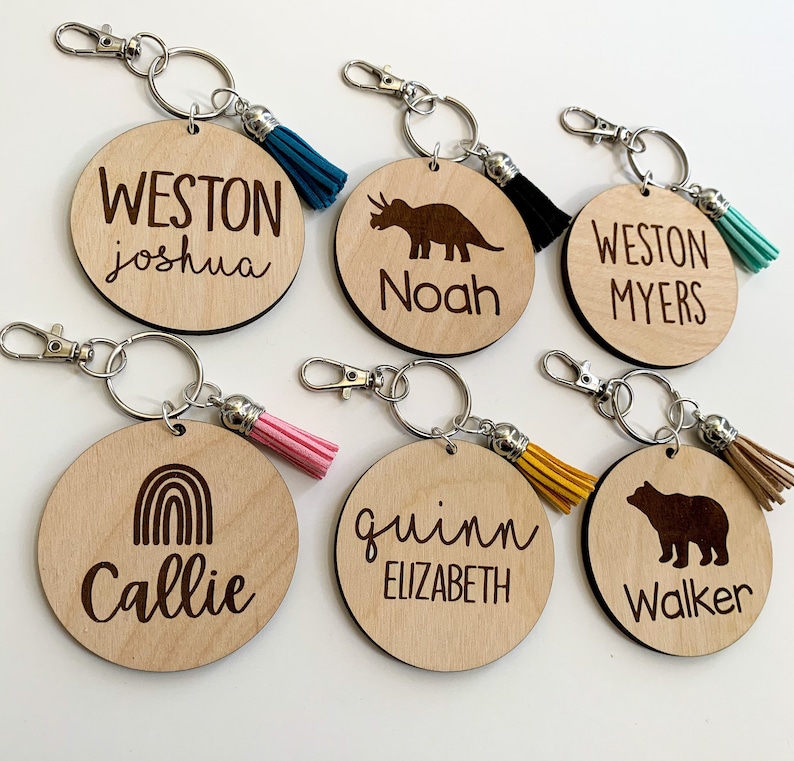May include: Six wooden keychains with different designs and names. The keychains feature a dinosaur, a rainbow, a bear, and the names Weston Joshua, Noah, Weston Myers, Callie, Quinn Elizabeth, and Walker. Each keychain has a small tassel.