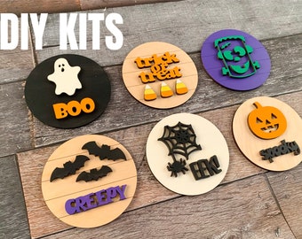 May include: Six DIY Halloween craft kits featuring various spooky themes. Each kit includes a round wooden base with a pre-cut design, including a ghost with "BOO", bats with "CREEPY", candy corn with "TRICK OR TREAT", a spider web with "EEK", a pumpkin with "SPOOKY", and a Frankenstein with a silly face.