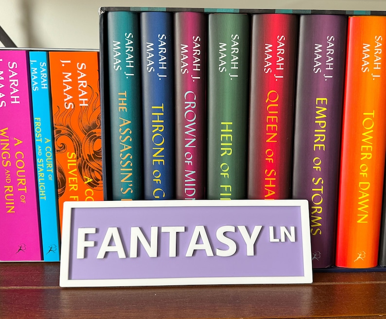 Peut inclure: Une &eacute;tag&egrave;re avec une pancarte violette indiquant "FANTASY LN" et une collection de romans fantastiques de Sarah J. Maas. Les livres sont rang&eacute;s dans l'ordre de publication, en commen&ccedil;ant par "Throne of Glass" et en terminant par "Tower of Dawn".