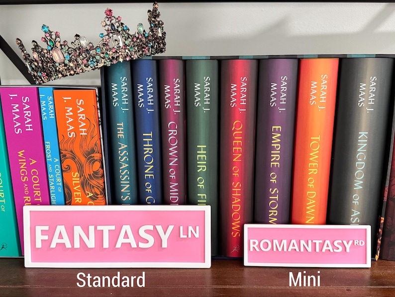 Peut inclure: Une &eacute;tag&egrave;re avec une collection de romans fantastiques de Sarah J. Maas. Les livres sont rang&eacute;s dans l'ordre de publication, en commen&ccedil;ant par "Throne of Glass" et en terminant par "Kingdom of Ash". Les livres sont tous en couverture rigide et ont un design similaire, avec un fond noir et des lettres dor&eacute;es. Il y a deux pancartes roses sur l'&eacute;tag&egrave;re, l'une qui dit "Fantasy LN" et l'autre qui dit "Romantasy RD".