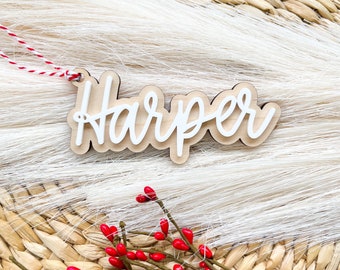 Op de afbeelding: Een houten kerstversiering met de naam "Harper" erop geschreven in witte verf. De versiering hangt aan een rood-wit koord en ligt op een wit, pluche oppervlak.