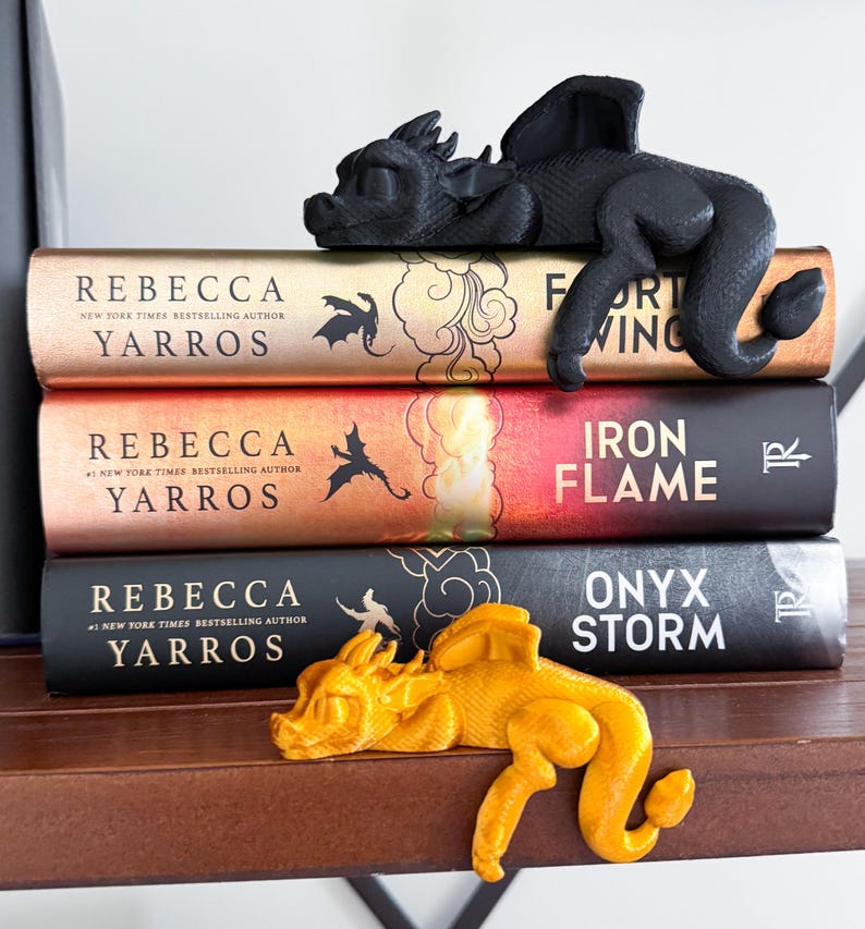May include: Three fantasy novels with dragon bookends. The top book is "Fort Wing" by Rebecca Yarros. The middle book is "Iron Flame" by Rebecca Yarros. The bottom book is "Onyx Storm" by Rebecca Yarros. The bookends are black and gold dragons.