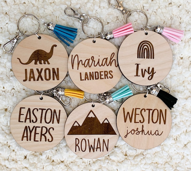 May include: Six wooden keychains with different designs and names. The keychains feature a dinosaur, a mountain range, a rainbow, and the names Jaxon, Mariah Landers, Ivy, Easton Ayers, Rowan, and Weston Joshua.