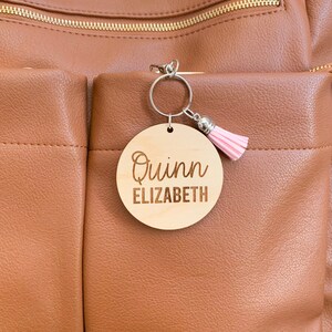 May include: A wooden keychain with the name "Quinn Elizabeth" engraved on it. The keychain is attached to a brown leather bag with a pink tassel.