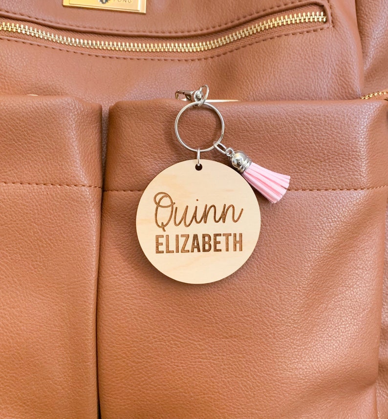 May include: A wooden keychain with the name "Quinn Elizabeth" engraved on it. The keychain is attached to a brown leather bag with a pink tassel.