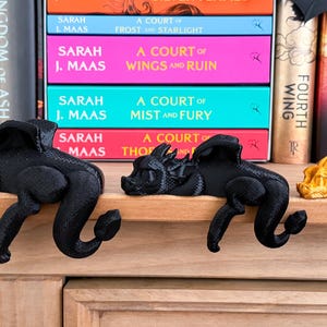 May include: Three 3D printed dragon bookends, one black, one black, and one gold, are perched on a wooden shelf. The bookends are in a sleeping position with their heads resting on the shelf. The shelf is in front of a bookshelf filled with books.