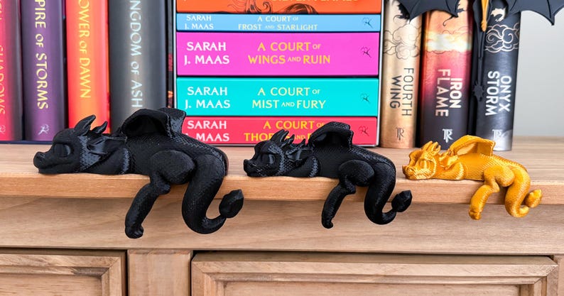 May include: Three 3D printed dragon bookends, one black, one black, and one gold, are perched on a wooden shelf. The bookends are in a sleeping position with their heads resting on the shelf. The shelf is in front of a bookshelf filled with books.
