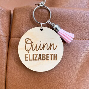 May include: A round wooden keychain with the name "Quinn Elizabeth" engraved on it. The keychain has a silver ring and a pink tassel.