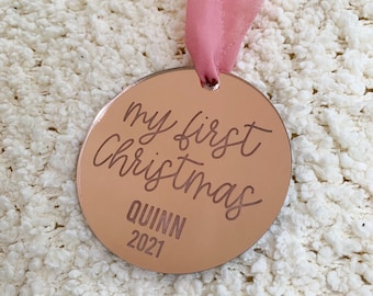 Puede incluir: Un adorno redondo de color oro rosa con el texto "my first christmas" y "QUINN 2021" en una fuente cursiva.