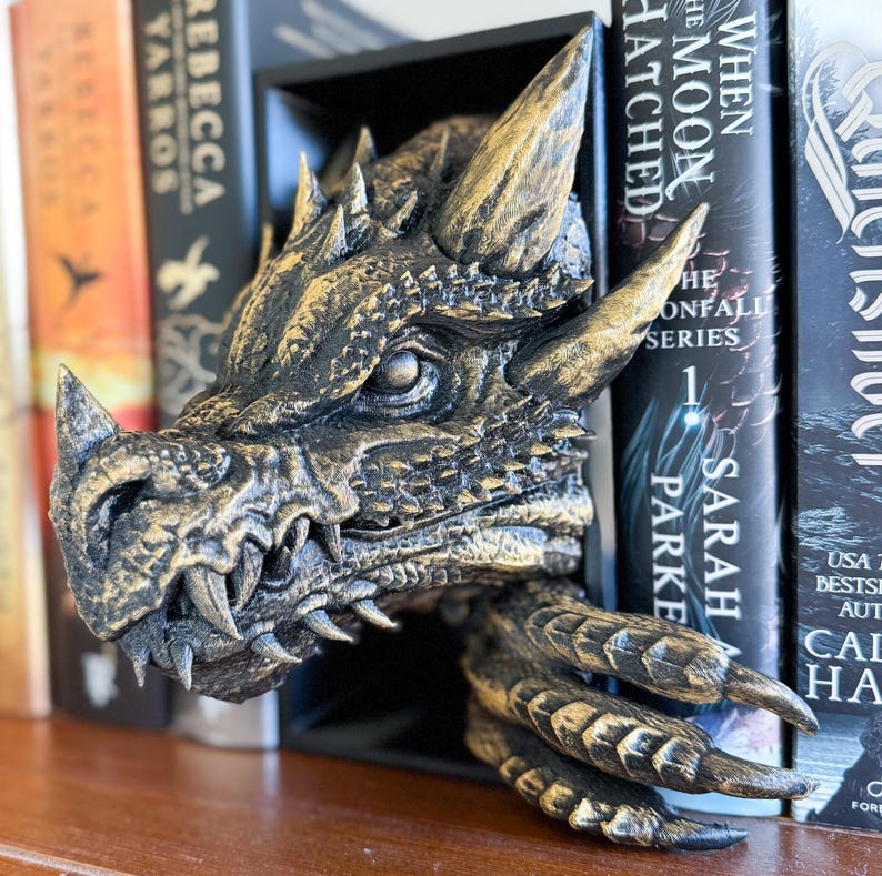 May include: A gold and black dragon head bookend with a textured surface. The dragon's mouth is open and its eyes are visible. The bookend is holding two books with the text "When the Moon Hatched" and "The Confall Series" visible.