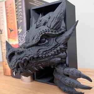 May include: Black dragon head bookend with sharp teeth and claws. The dragon head is mounted in a black frame and is a 3D printed sculpture.