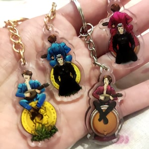 May include: Keychains featuring a musician playing a stringed instrument. The musician is depicted in various outfits, sitting on a coin, a barrel, or a drum. The keychains have gold or silver chains and rings.