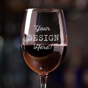 May include: A clear wine glass filled with red wine, resting on a wooden surface. The glass has the text "Your DESIGN Here!" etched on the side. The background is blurred, suggesting a bar setting.