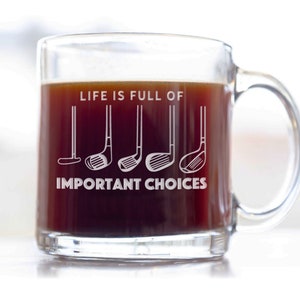 May include: A clear glass coffee mug with a black handle. The mug has the text "Life is full of" and a row of five golf clubs with the text "Important Choices" below.