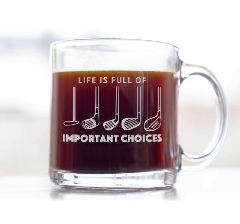 May include: A clear glass coffee mug with a black handle. The mug has the text "Life is full of" and a row of five golf clubs with the text "Important Choices" below.