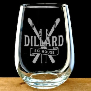 May include: A clear glass stemless wine glass with an etched design of crossed skis and the text "Dillard Ski House".