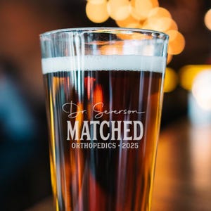 May include: A clear glass beer stein with the text "Dr. Severson MATCHED ORTHOPEDICS - 2025" etched on the side. The stein is filled with a dark brown beer.