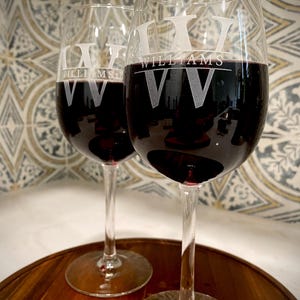 May include: Two clear glass wine glasses with red wine. The glasses are engraved with the name "Williams" and the initials "VV".