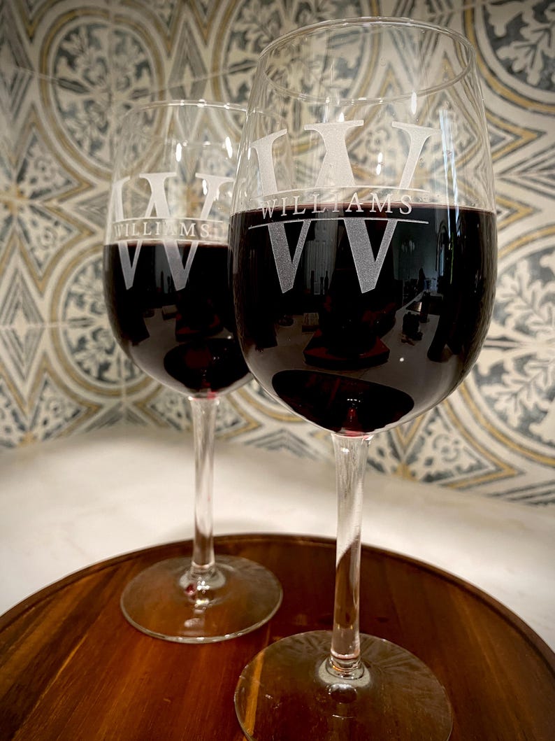 May include: Two clear glass wine glasses with red wine. The glasses are engraved with the name "Williams" and the initials "VV".