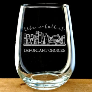 May include: A clear glass stemless wine glass with the text "life is full of IMPORTANT CHOICES" and an illustration of a bookshelf full of books.