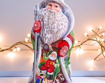 May include: A hand-painted Santa Claus figurine. The figure wears a white hat and coat with red mittens. The figurine holds a silver staff and features a painted scene of Santa relaxing by a fireplace with a dog and presents.