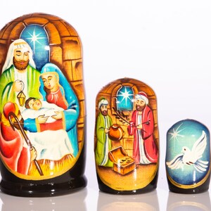 May include: A set of three wooden nesting dolls depicting the Nativity scene. The largest doll shows Mary, Joseph, and the baby Jesus in a stable. The middle doll shows two wise men bringing gifts. The smallest doll shows a white dove.
