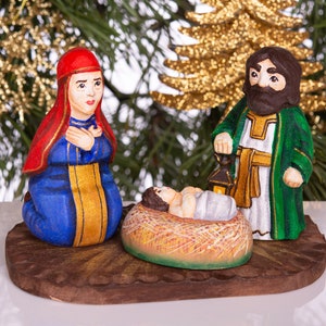 May include: A wooden nativity scene with a red-robed Mary kneeling beside a manger with a baby Jesus inside. Joseph is standing behind the manger, wearing a green robe and holding a lantern.