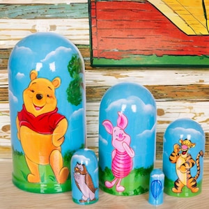 May include: A set of five Winnie the Pooh-themed nesting dolls. The dolls feature illustrations of Winnie the Pooh, Piglet, Tigger, Owl, and a blue balloon. Each doll is painted with a blue sky background and green grass.