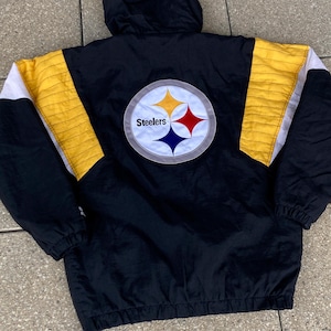 Könnte beinhalten: Schwarze Kapuzenjacke mit gelben und weißen Akzenten. Auf der Rückseite befindet sich ein kreisförmiges Steelers-Logo mit einem gelben, roten und blauen Sternmuster. Die Jacke hat eine gesteppte gelbe Schulterpartie und weiße Streifen.