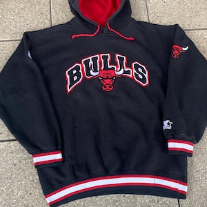 Könnte beinhalten: Schwarzer Chicago Bulls Kapuzenpullover mit roter Kapuzeninnenseite. Das Wort "BULLS" steht in Weiß mit roten Akzenten auf der Brust, und ein Bullenlogo befindet sich in der Mitte. Die Ärmel und der Saum haben weiße und rote Streifen.
