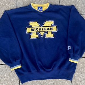 Peut inclure: Un sweat-shirt bleu marine avec le logo "MICHIGAN" jaune et blanc. Le col et les poignets sont ornés de garnitures jaunes et grises. Le logo Starter est visible sur la manche.