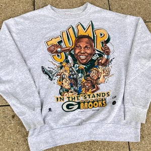Könnte beinhalten: Hellgraues Sweatshirt mit farbenfroher Grafik eines Footballspielers und dem Text "JUMP IN THE STANDS BROOKS". Die Grafik enthält das Logo der Green Bay Packers und Cartoonfiguren. Das Sweatshirt hat einen Rundhalsausschnitt und lange Ärmel.