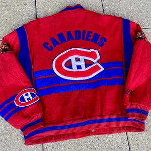 Op de afbeelding: Rode en blauwe vintage jas met het woord "CANADIENS" over de rug. De jas heeft een groot "C"-logo met een "H" erin en blauwe strepen op de mouwen. Het NHL-logo staat op de mouw.