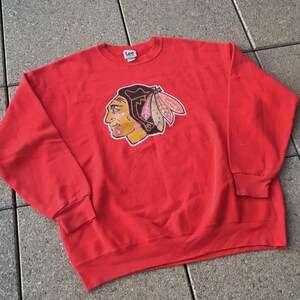 Könnte beinhalten: Ein rotes Sweatshirt mit Rundhalsausschnitt und einer Grafik eines indianischen Häuptlings im Profil. Die Grafik enthält gelbe, schwarze und rosa Details. Das Sweatshirt hat lange Ärmel und gerippte Bündchen und Saum. Die Marke "Lee" ist sichtbar.