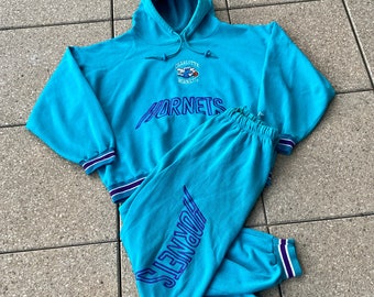 May include: A turquoise tracksuit comprising a hooded top and matching trousers. The top and trousers feature the word "HORNETS" in blue, with a logo on the top. Both items have striped cuffs and hems.