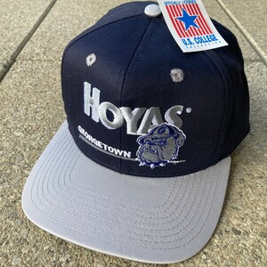 Peut inclure: Une casquette de baseball bleu marine et grise avec le mot "Hoyas" et "Georgetown" brodé en argent sur le devant. Un bouledogue argenté mascotte est également brodé sur le devant de la casquette. La casquette a une visière grise et une étiquette blanche qui dit "Officially Licensed U.S. College Collection" attachée sur le côté.