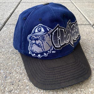 Peut inclure: Une casquette de baseball bleue et noire avec un mascotte de bouledogue et le mot "Huskies" dans une police de caractères stylisée.