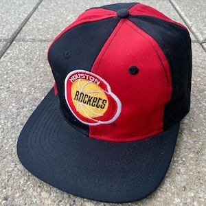 Op de afbeelding: Een zwart en rode Houston Rockets baseball cap. De cap heeft het teamlogo op de voorkant, een basketbal met de tekst "Houston Rockets" in een cirkelvormig ontwerp. De cap is gemaakt van een duurzame stof.