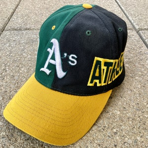 Könnte beinhalten: Baseballkappe in Grün, Schwarz und Gold. Auf der Kappe sind die Buchstaben "A's" in Weiß auf der Vorderseite und das Wort "ATHLETICS" in Gelb auf der Seite zu sehen. Der Schirm ist goldfarben.